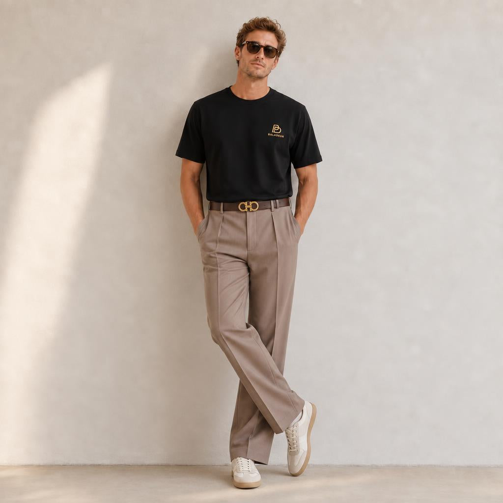 A man wearing a black Baladeur short-sleeve t-shirt with a small gold logo on the left chest, paired with beige pleated trousers, a brown belt with a gold buckle, and white sneakers, standing against a textured light gray wall.