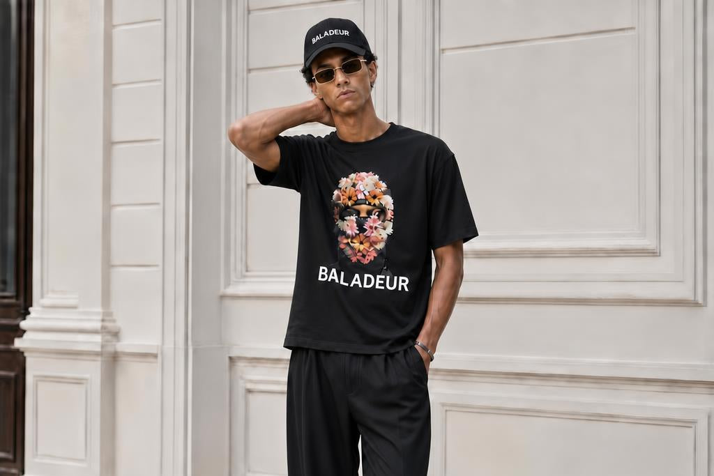 A man wearing a black Baladeur t-shirt with a floral graphic and matching black Baladeur pants, standing in front of a white paneled building wall.