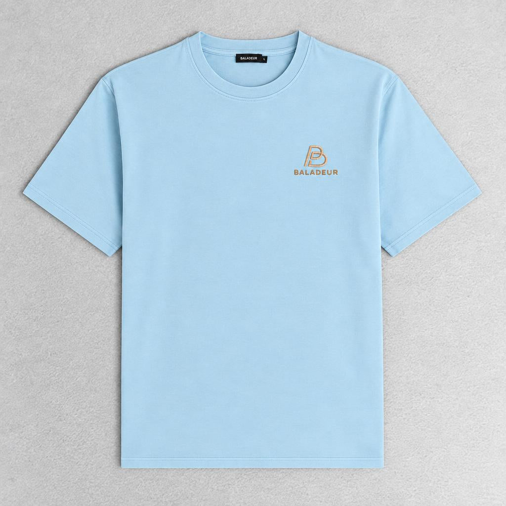 A light blue short-sleeve crewneck t-shirt with a small gold Baladeur logo on the left chest, laid flat on a textured gray background.