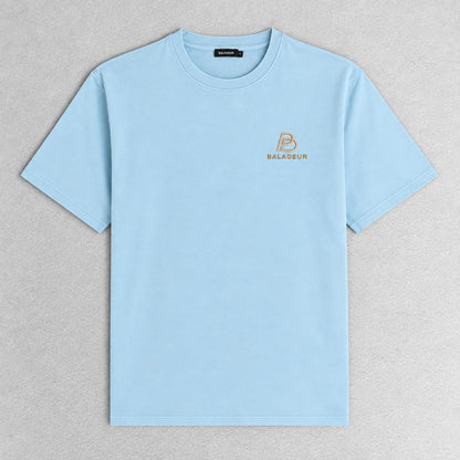 A light blue short-sleeve crewneck t-shirt with a small gold Baladeur logo on the left chest, laid flat on a textured gray background.