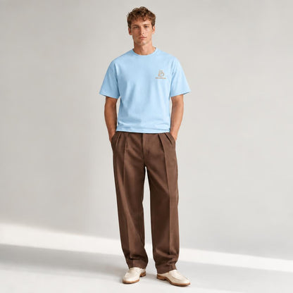 A man modeling a light blue BALADEUR crewneck t-shirt with a small embroidered logo on the left chest, paired with brown pleated trousers and cream-colored loafers, set against a neutral light gray studio background.