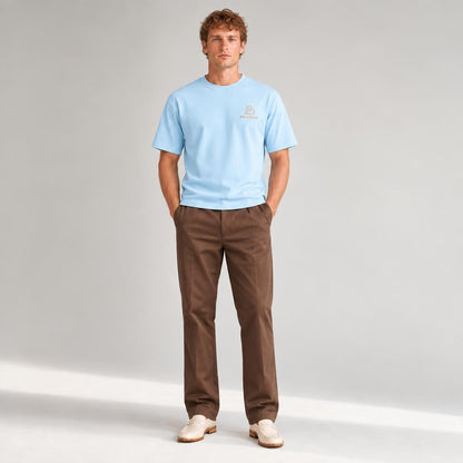 A man stands against a light gray studio background wearing a light blue crew-neck t-shirt with a small BALADEUR embroidered logo on the left chest, brown trousers, and off-white loafers.