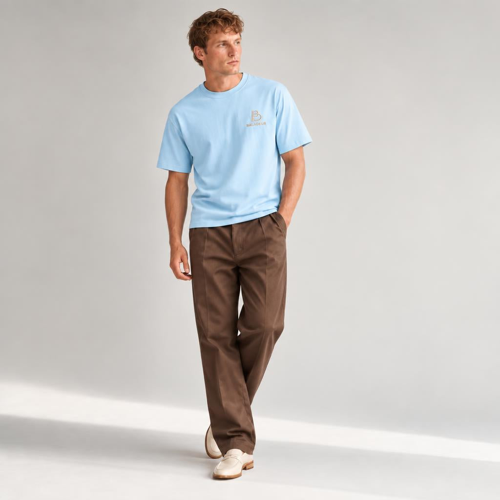 A man modeling a light blue crewneck t-shirt with a small embroidered  BALADEUR logo on the left chest, paired with brown trousers and off-white sneakers, set against a plain light gray studio background.