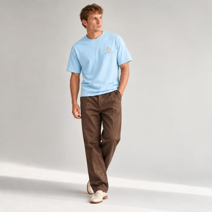 A man modeling a light blue crewneck t-shirt with a small embroidered  BALADEUR logo on the left chest, paired with brown trousers and off-white sneakers, set against a plain light gray studio background.