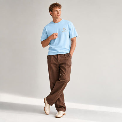 A man modeling a light blue crewneck t-shirt with a small gold-beige BALADEUR embroidered logo on the left chest, paired with brown trousers and cream-colored shoes, standing against a light gray studio background.