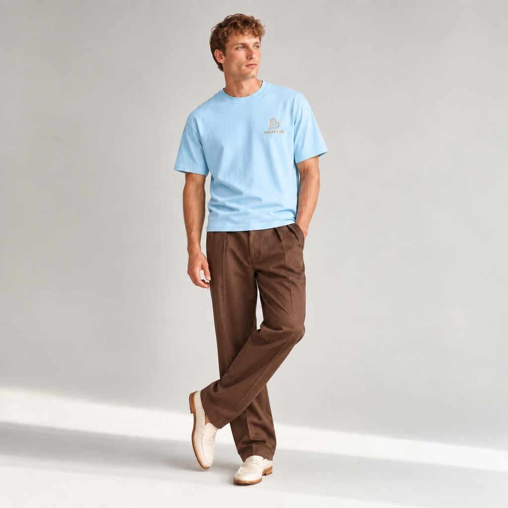 A man wearing a light blue crewneck t-shirt with a small BALADEUR embroidered logo on the left chest, paired with brown trousers and cream-colored loafers, standing against a light gray studio background.