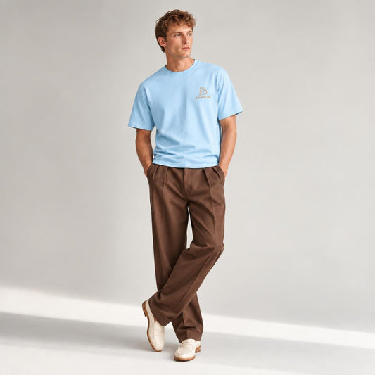 A man wearing a light blue crewneck t-shirt with a small BALADEUR embroidered logo on the left chest, paired with brown pleated trousers and off-white loafers, standing against a light gray studio background.
