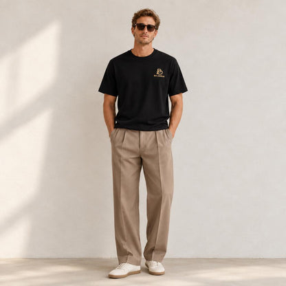 A man wearing a black short-sleeve BALADEUR t-shirt with a small gold logo on the left chest, paired with beige pleated trousers and white sneakers, standing against a light gray wall with soft shadows.