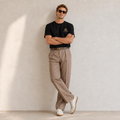 A man wearing a black crew-neck BALADEUR t-shirt with a small gold logo on the left chest, beige pleated trousers, white sneakers, and sunglasses, leaning against a textured light gray wall.