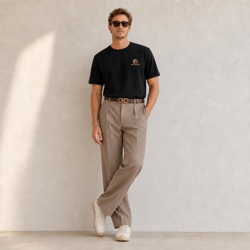 A man wearing a black short-sleeve BALADEUR t-shirt with a small gold logo on the left chest, beige trousers, a brown belt with a gold buckle, and white sneakers, standing against a textured light gray wall.
