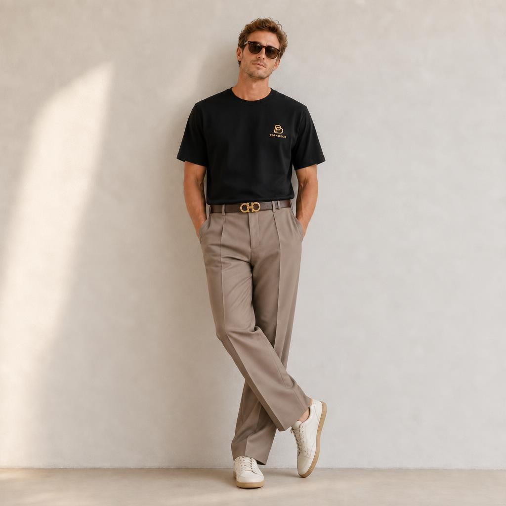A man wearing a black short-sleeve BALADEUR t-shirt with a small brown-beige logo on the left chest, paired with beige trousers, a brown belt with a gold buckle, and white sneakers, leaning against a textured light gray wall.