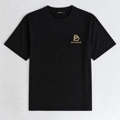 A black short-sleeve crewneck t-shirt with a small gold Baladeur logo printed on the left chest, displayed flat against a light gray textured background.