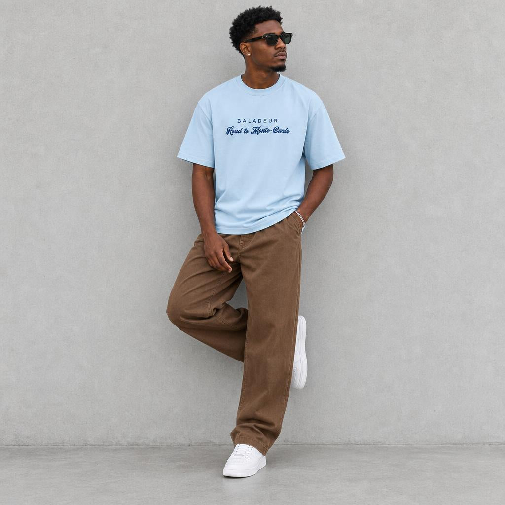 A man wearing a light blue t-shirt with 'BALADEUR Road to Monte Carlo' by 'BALADEUR' printed on the chest, paired with brown pants and white sneakers, posing against a textured grey wall.