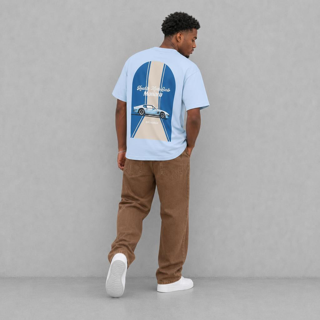 A man wearing a light blue t-shirt with a large graphic print on the back, paired with brown corduroy pants and white sneakers, standing against a gray studio background. The brand name is BALADEUR 