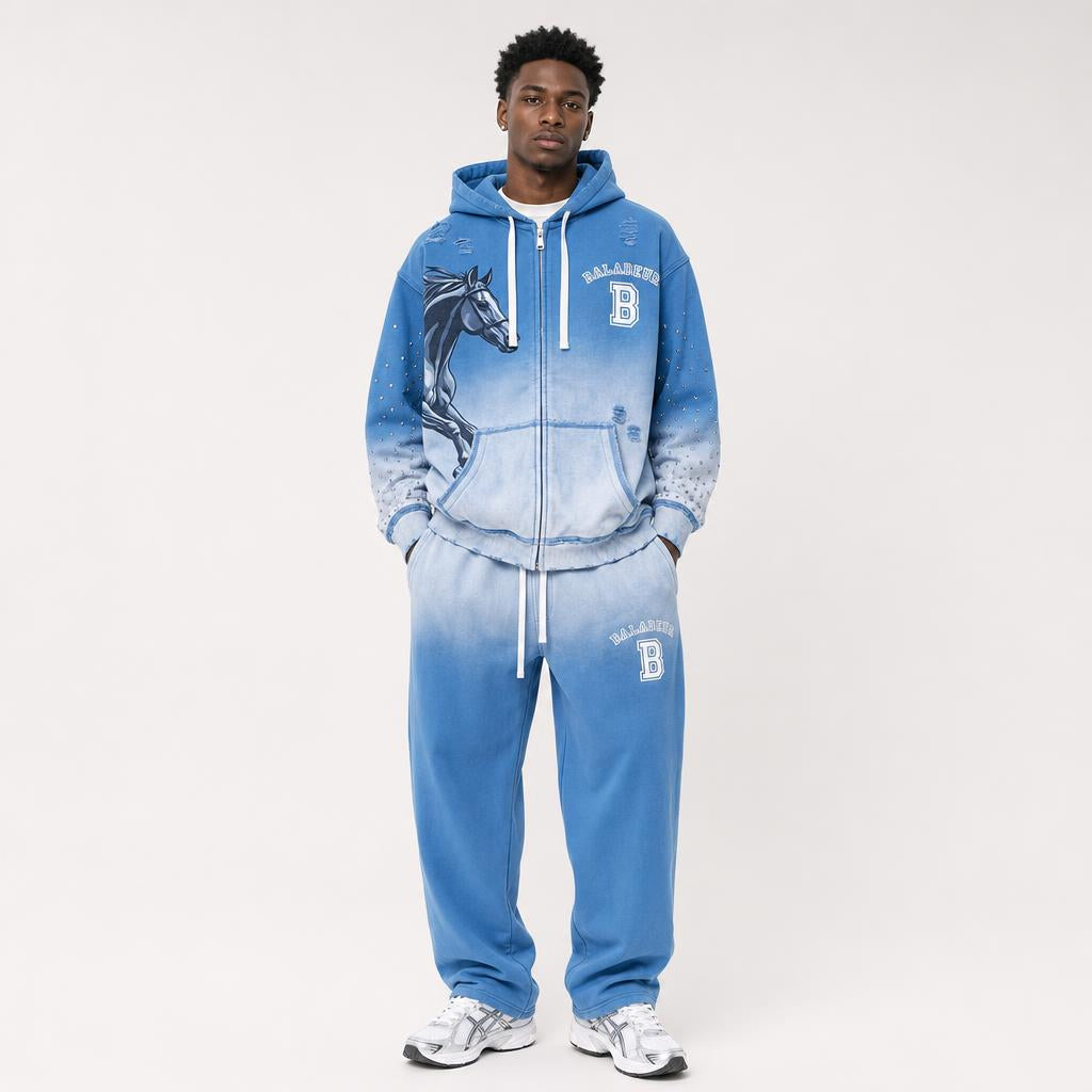 A man modeling a blue zip-up hoodie and matching sweatpants set with a white gradient fade, featuring a horse graphic on the hoodie, white drawstrings, and 'BALADEUR' text with a 'B' logo on the front and thigh.