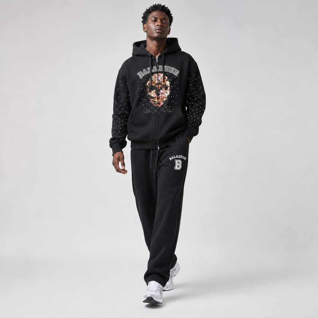 A man modeling a black zip-up hoodie and matching sweatpants set from Baladeur, featuring a skull graphic on the chest with a gold skull and white brand lettering, and a white 'B' logo on the thigh.