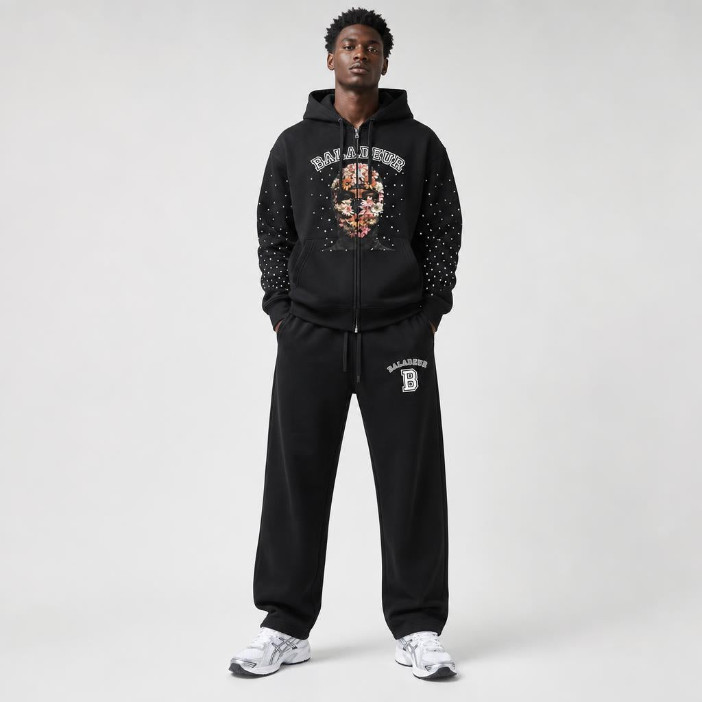 A male model wearing a black BALADEUR zip-up hoodie and matching sweatpants with white sneakers, standing against a light gray studio background.