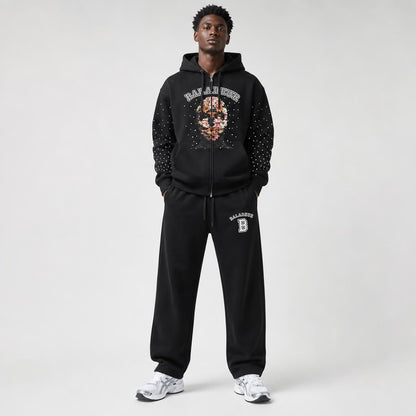 A male model wearing a black BALADEUR zip-up hoodie and matching sweatpants with white sneakers, standing against a light gray studio background.