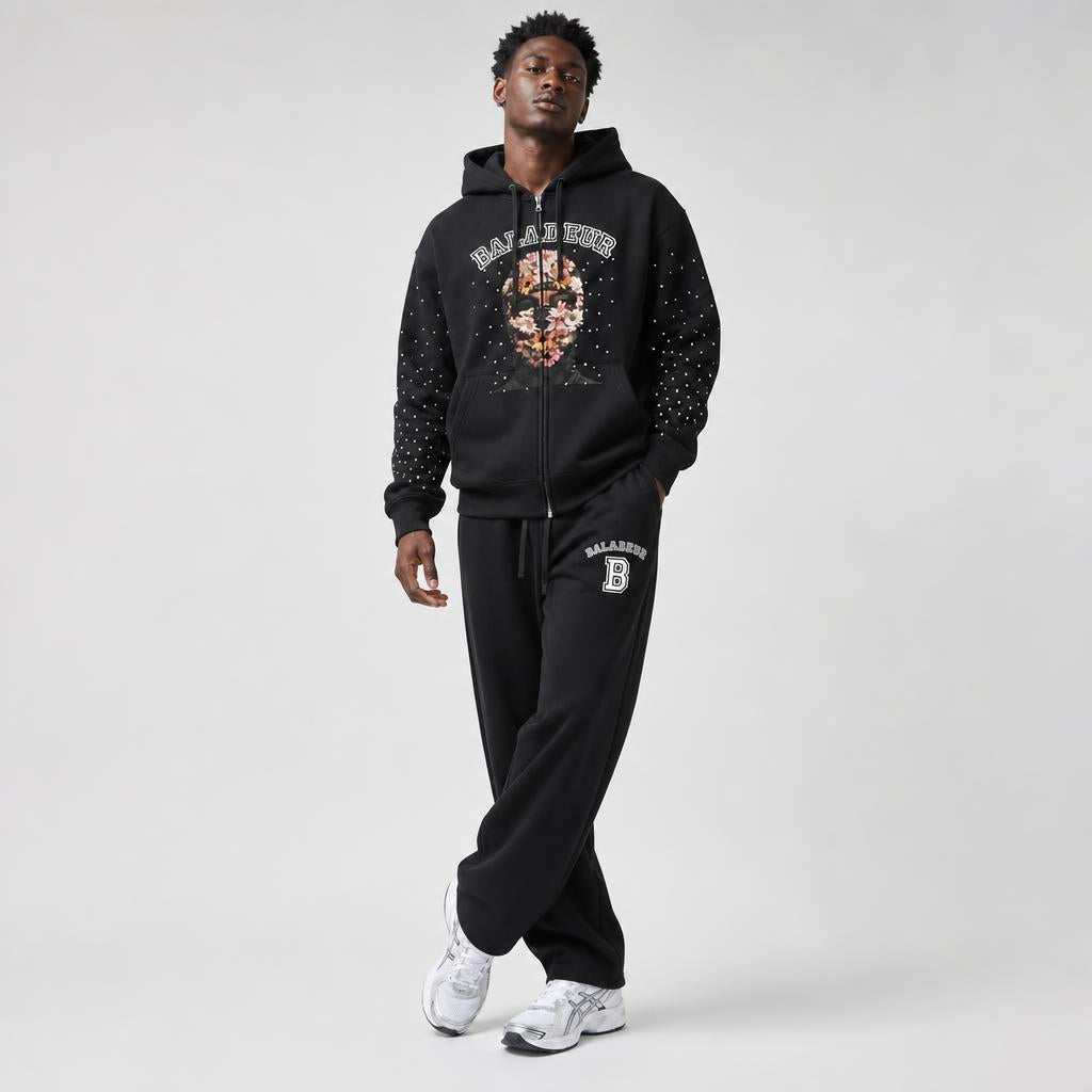 A man modeling a black zip-up hoodie and matching sweatpants set with 'BALADEUR' graphics, including a floral pattern on the hoodie chest and a white 'B' logo on the thigh, against a light gray studio background.