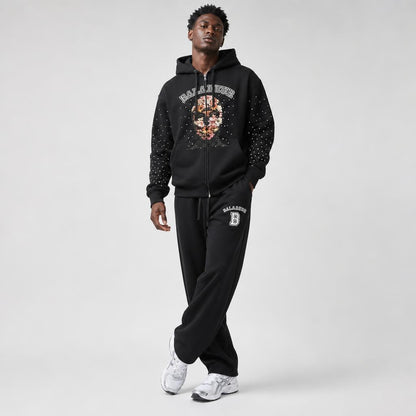 A man modeling a black zip-up hoodie and matching sweatpants set with 'BALADEUR' graphics, including a floral pattern on the hoodie chest and a white 'B' logo on the thigh, against a light gray studio background.