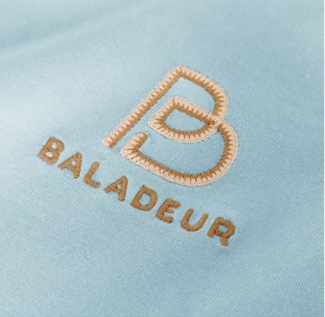 Close-up of a light blue fabric surface with an embroidered brown-beige monogram logo and the word BALADEUR stitched below it.