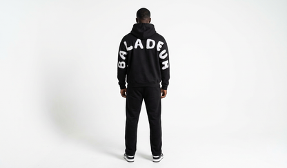 Person wearing a black tracksuit with 'Baladeur' branding on a white background